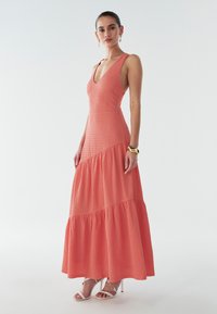 Koralowa, bezrękawna maxi sukienka z dekoltem w kształcie litery V, o teksturowanej powierzchni, z falbaniastego projektu spódnicy, noszona z białymi sandałami na obcasie i złotymi dodatkami.