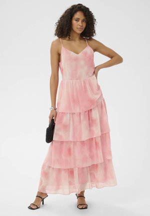 Žena s kudrnatými vlasy, oblečená v růžových vícevrstvých maxišatech bez rukávů, drží černou kabelku a stojí s jednou rukou v bok.
