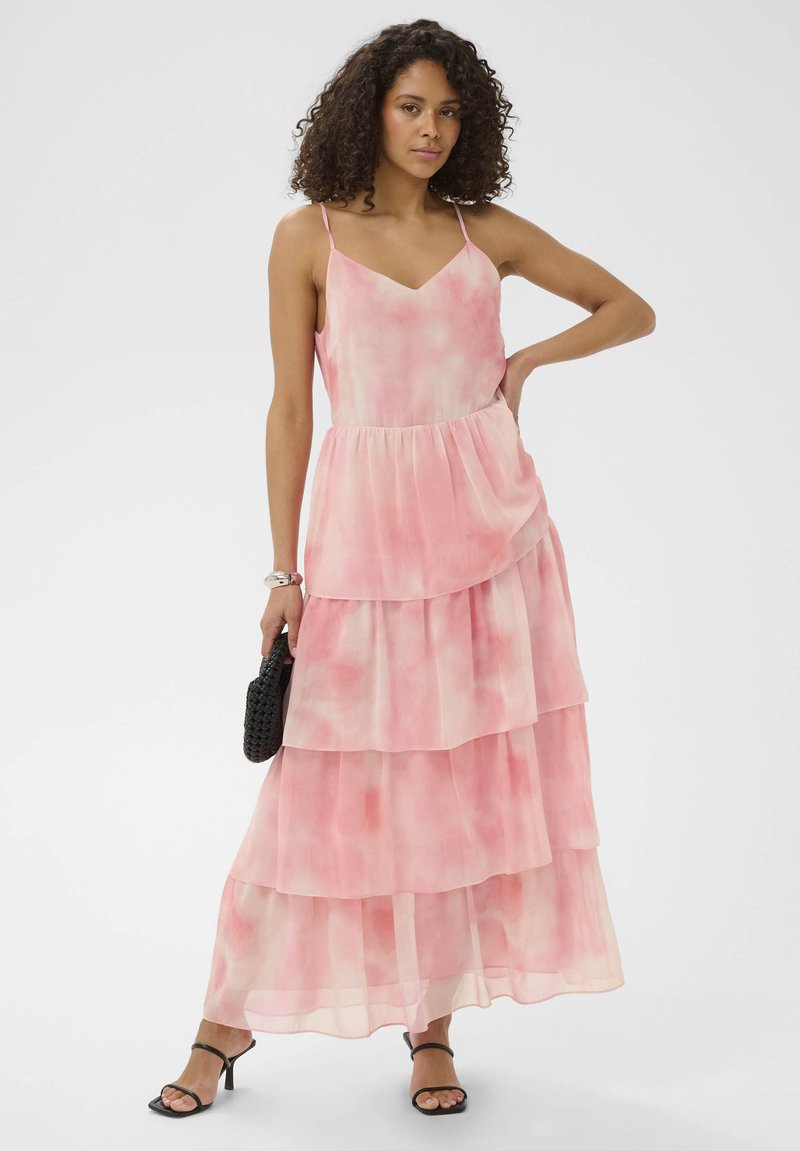 Femme aux cheveux bouclés portant une robe longue rose sans manches à volants, tenant un sac noir, debout avec une main sur la hanche.