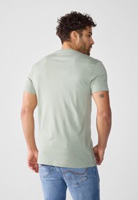 Camiseta de manga corta verde claro con cuello redondo, textura suave y corte ligeramente ajustado. Presenta un tatuaje visible en el brazo izquierdo.