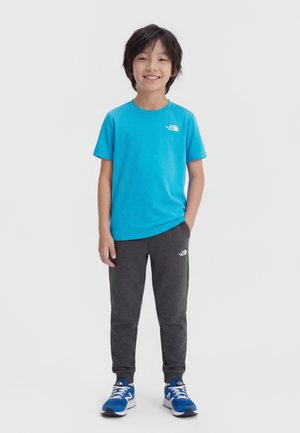 Mosolygó fiú világoskék pólóban, sötétszürke melegítőnadrágban és kék tornacipőben, egy egyszerű fehér háttér előtt áll.