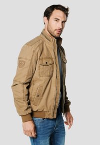 Béžová bomber bunda s stojáčkovým límcem, zapínáním na zip, dvěma předními kapsami a žebrovanými manžetami. Vyznačuje se strukturovanou látkou a nášivkou na rukávu.