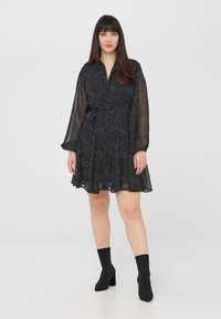 Abito nero a pois con design avvolgente, maniche lunghe trasparenti e gonna svasata. Abbinato a stivaletti neri alla caviglia.