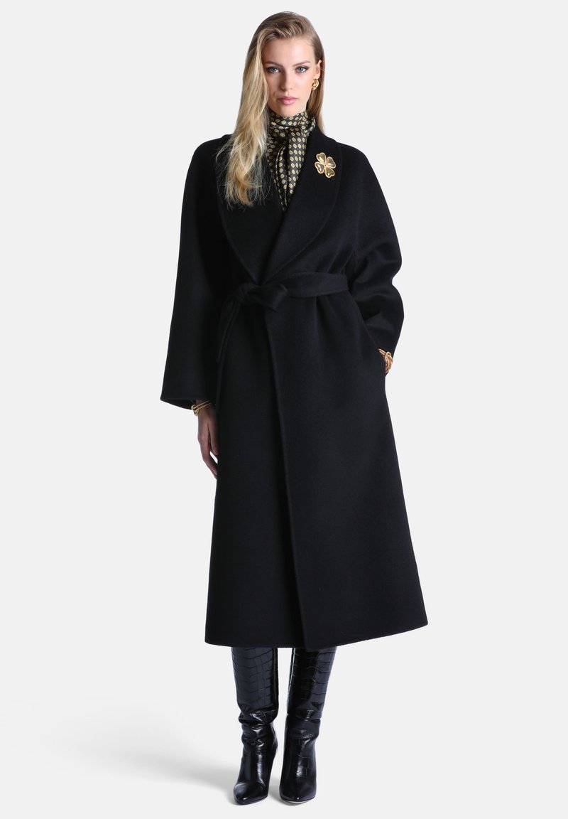 Femme portant un long manteau noir ceinturé, une blouse à motifs à col montant, des bottes noires hautes jusqu'au genou, avec une broche fleur dorée et des boucles d'oreilles.