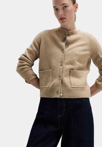 Beżowy kardigan wełniany z okrągłym dekoltem, prążkowanym dołem i mankietami, dwoma przednimi kieszeniami oraz zapięciem na guziki z dekoracyjnymi guzikami.