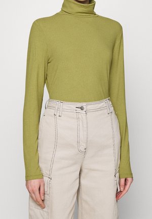 Henkilö pukeutuneena ribbineulottuun oliivinvihreään poolopaitaan ja korkeavyötäröisiin beigenvärisiin housuihin, joissa on näkyvät saumat ja sivutaskut.