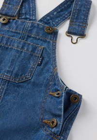 Blauwe denim overall met verstelbare banden, messing beslag, gestikte accenten en een label op de voorzak. Textuur is klassieke denim.