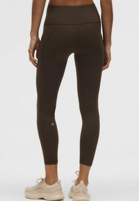 Tagumine vaade inimesest, kes kannab tumedast pruunist kangast legginsit koos küljetaskuga ja beeže jooksujalatseid tähnilise tausta ees.