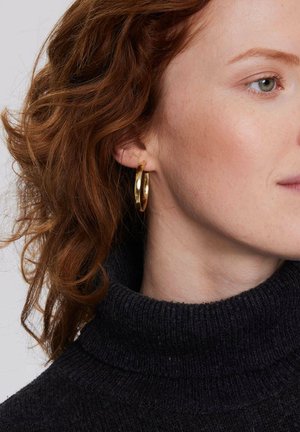 Złota kolczyk w kształcie obręczy o polerowanej powierzchni, okrągłym kształcie i gładkiej teksturze, noszony przez modelkę z włosami średniej długości w falach.