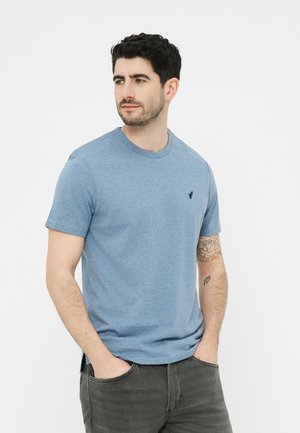 Uomo con capelli scuri che indossa una maglietta blu a maniche corte e jeans grigi, in piedi con le mani nelle tasche, guarda a sinistra.