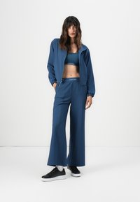 Mørkeblå treningstøy sett som inkluderer en jakke med glidelås, en matchende crop top, og vide bukser. Svart sneakers fullfører stilen.