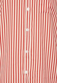 Chemise rayée rouge et crème avec des lignes verticales, dotée de boutons blancs sur le devant et d'une poche poitrine. Texture de tissu lisse.