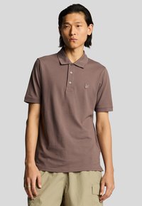 Polo de color marrón con cuello, que presenta tres botones y un pequeño logo bordado en el lado izquierdo del pecho. Fabricado con una tela suave.