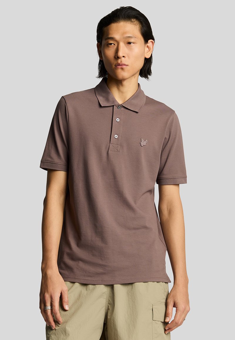 Polo de color marrón con cuello, que presenta tres botones y un pequeño logo bordado en el lado izquierdo del pecho. Fabricado con una tela suave.