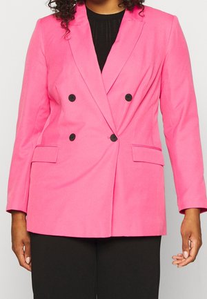 Woman wearing bright pink double-breasted blazer with black buttons over a black top and black pants, standing against a plain background.