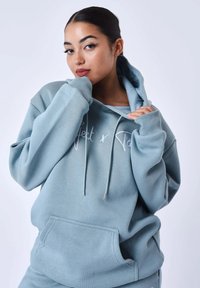 Sweatshirt à capuche bleu clair avec une poche avant, des cordons de serrage et une écriture blanche. Tissu doux et design à coupe ample.