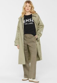 Hellgrüner langer Mantel über einem schwarzen Sweatshirt mit weißem Text, kombiniert mit karierten weitgeschnittenen Hosen und beigen Sneakers.