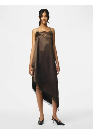 Femme portant une robe asymétrique en satin marron foncé sans manches avec une bordure en dentelle et des talons slingback noirs pointus, debout devant un fond blanc.