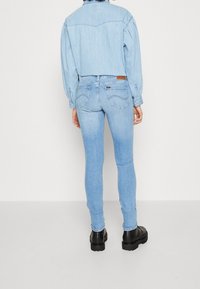 Jeans de mezclilla azul claro con un corte slim, que presentan una cintura alta, un ligero desgaste y detalles de costura en los bolsillos traseros. Combinados con una camisa de mezclilla azul claro.