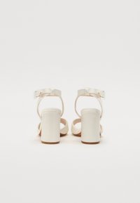Λευκές ψηλές σάνταλες με φαρδύ τακούνι, ρυθμιζόμενο λουράκι στον αστράγαλο και διακοσμητική λεπτομέρεια πίσω. Λείος άνω υλικό και υφασμάτινα αξεσουάρ.