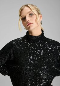 Suéter negro con lentejuelas, de cuello alto, corte relajado y superficie texturizada, que refleja la luz con un acabado brillante.