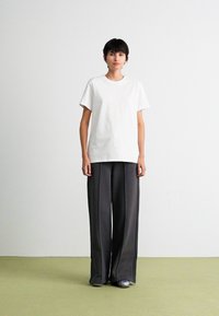 Personne se tenant contre un mur blanc, portant un T-shirt blanc à manches courtes, un pantalon large gris foncé ample et des chaussures grises sur un sol vert.