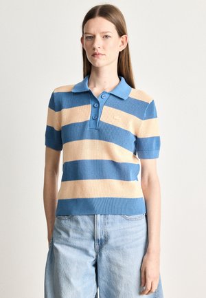 Donna che indossa una polo a maniche corte a righe blu e beige e jeans in denim chiaro a vita alta, in piedi su uno sfondo neutro.