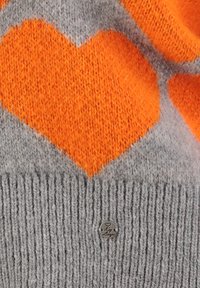 Nahaufnahme eines grauen Strickstoffs mit einem orangefarbenen Herzmuster und einem kleinen runden Metallschild, das nahe dem unteren Ende befestigt ist.