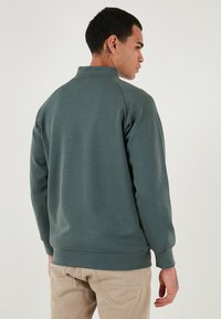 Sudadera de color verde oscuro con cuello alto y puños acanalados, fabricada en un tejido suave; mostrada desde un ángulo trasero.