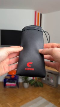Estuche para gafas Carrera negro con textura suave, cierre de cremallera y un logo rojo. Forma rectangular, sostenido por dos manos.