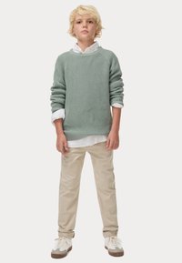 Maglione a maglia verde chiaro con texture a coste, indossato sopra una camicia bianca con colletto. Pantaloni beige e sneakers bianche con suole marroni.