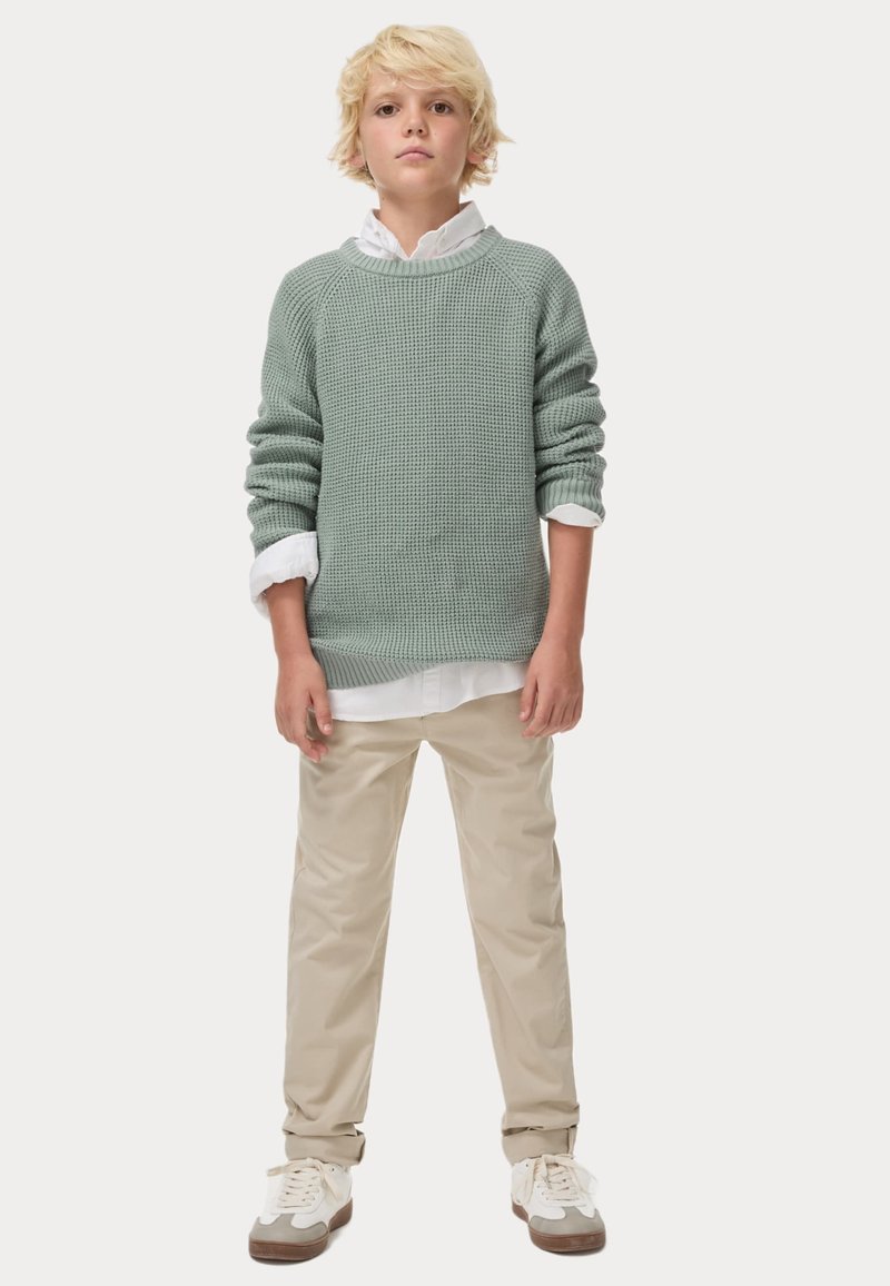 Maglione a maglia verde chiaro con texture a coste, indossato sopra una camicia bianca con colletto. Pantaloni beige e sneakers bianche con suole marroni.