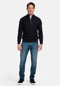 Schwarzer Strickpullover mit einem Hals-Reißverschluss, kombiniert mit blauen Jeans und schwarzen Sneakers. Hat eine gerippte Textur und eine taillierte Passform.
