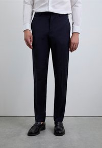 Pantalones azul marino hechos de tela lisa, con un corte ajustado, líneas limpias y bolsillos; combinados con mocasines negros sobre una superficie gris.