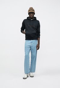 Jauns vīrietis, kas valkā svītrainu cepuri, melnu Jack & Jones kapuci, gaiši zilo platajā zeķēs un baltas kedas ar melnām svītrām.