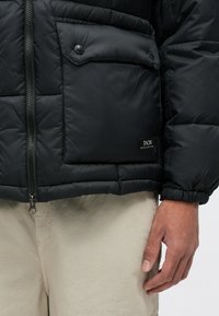 Μαύρο μπουφάν από πούπουλο με καπιτονέ σχέδιο, διαθέτοντας κλείσιμο με φερμουάρ μπροστά και τσέπη με καπάκι και κουμπιά. Ελαστικοί καρποί και ετικέτα μάρκας ορατοί.