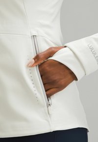 Jachetă albă ajustată cu fermoar lateral și buzunar pentru mâini. Textură netedă, detalii reflectorizante și găuri ascunse pentru degete pe mâneci.