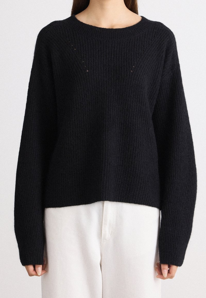Femme portant un pull en maille côtelée noir à manches longues avec un col rond et un pantalon blanc, vue des épaules jusqu'aux hanches.