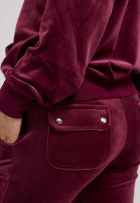Burgundy velvet joggers with a side pocket, featuring shiny silver hardware. The fabric has a soft, smooth texture and a fitted design.