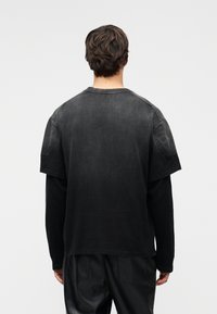 Μαύρο υπερμεγέθες μπλουζάκι με ξεθωριασμένη υφή, κοντά μανίκια και στρογγυλή λαιμόκοψη. Η πίσω πλευρά έχει λεία επιφάνεια και διακριτικές ραφές.