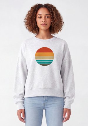 Jeune femme aux cheveux bouclés portant un sweat-shirt gris clair avec un cercle de style rétro composé de bandes horizontales colorées dans des tons terre.
