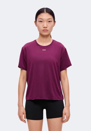 Une femme pose avec un haut de sport à manches courtes couleur prune et un short noir sur un fond clair et uni.