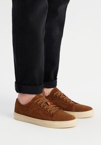 Baskets en daim marron avec bout arrondi, semelle en caoutchouc crème et lacets. Présentent un petit accent multicolore près des œillets. Portées avec un jean noir.