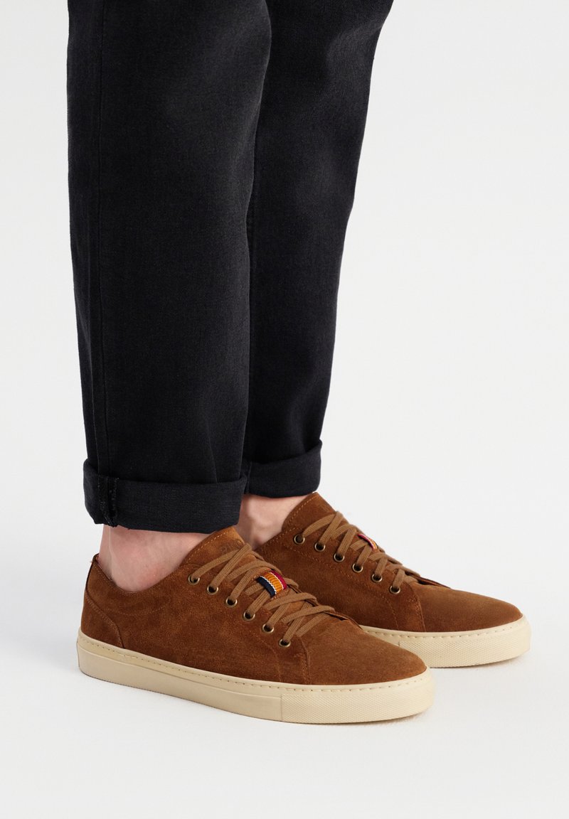 Adidași maro din suede, cu vârf rotund, talpă din cauciuc crem și șireturi. Au un mic accent multicolor lângă ochiurile de șiret. Purtați cu blugi negri.