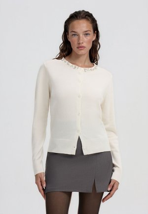 Donna con capelli effetto bagnato che indossa un cardigan bianco con bottoni e dettagli di perle e una gonna grigia con uno spacco frontale, in piedi contro uno sfondo semplice.