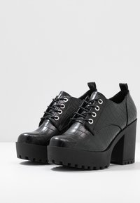 Zapatos de plataforma negros con una textura de cocodrilo brillante, puntera redonda, parte delantera con cordones y tacones gruesos de bloque con una suela estriada.