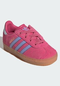 Sapatilha de camurça rosa com três riscas azul-claro, sola de borracha texturizada e atacadores rosa a condizer. Apresenta uma aba de calcanhar prateada e marcação.