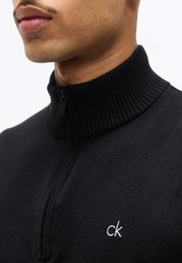 Κοντινή λήψη ενός ατόμου που φοράει μαύρο πουλόβερ με φερμουάρ, ριγέ γιακά και λευκό λογότυπο «ck» στο στήθος.