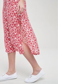 Robe à motif floral rouge avec des fentes, faite d'un tissu léger. Associée à des baskets blanches avec des boutons dorés.