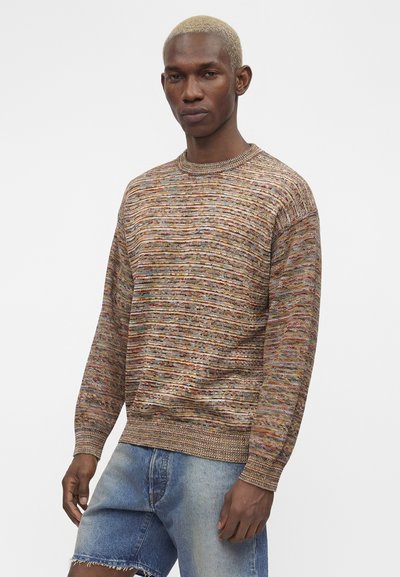 Άνδρας με κοντά ξανθά μαλλιά που φοράει πολύχρωμο πλεκτό πουλόβερ και ανοιχτό μπλε τζιν σορτς σε απλό λευκό φόντο.